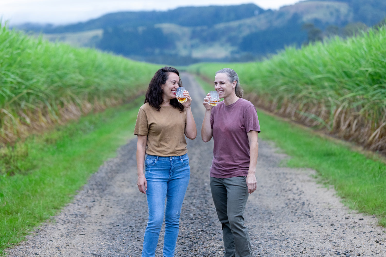 Two women in sugarcane fields with glasses of rum.